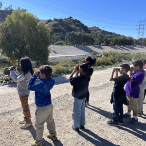 Students use binoculars to look at the LA River on a sunny day.