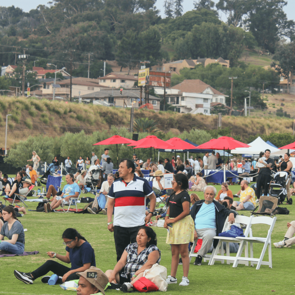 River Fest 2023 attendees sit on blankets and chairs at a large outdoor grass area at Los Angeles State Historic Park as they watch a performance on stage .