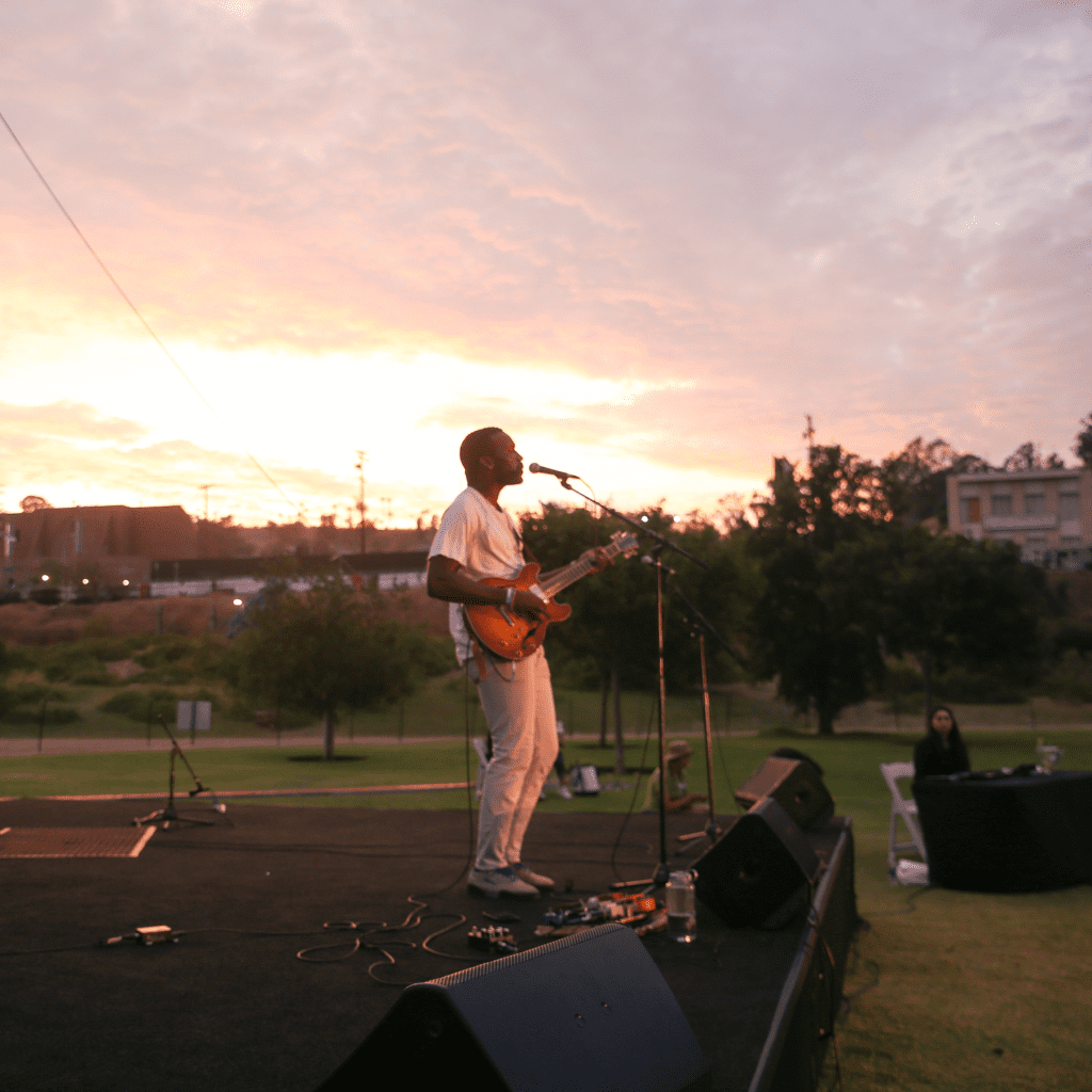 Christopher Watson sings and plays electric guitar on stage as the sun sets behind him for Friends of the Los Angeles River's 2023 River Fest event at Los Angeles State Historic Park.