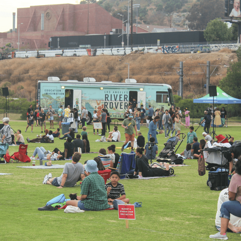River Fest 2023 attendees sit on blankets and walk around a large grass area at Los Angeles State Historic Park, with the River Rover parked in the distance. The River Rover is Friends of the Los Angeles River's thirty-eight foot long recreational vehicle that serves as a mobile museum and education center.
