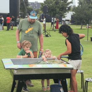 A FoLAR Educator pours water over FoLAR's three-dimensional model of the Los Angeles River Watershed, teaching two young children and their guardian about watershed flows at River Fest 2023.